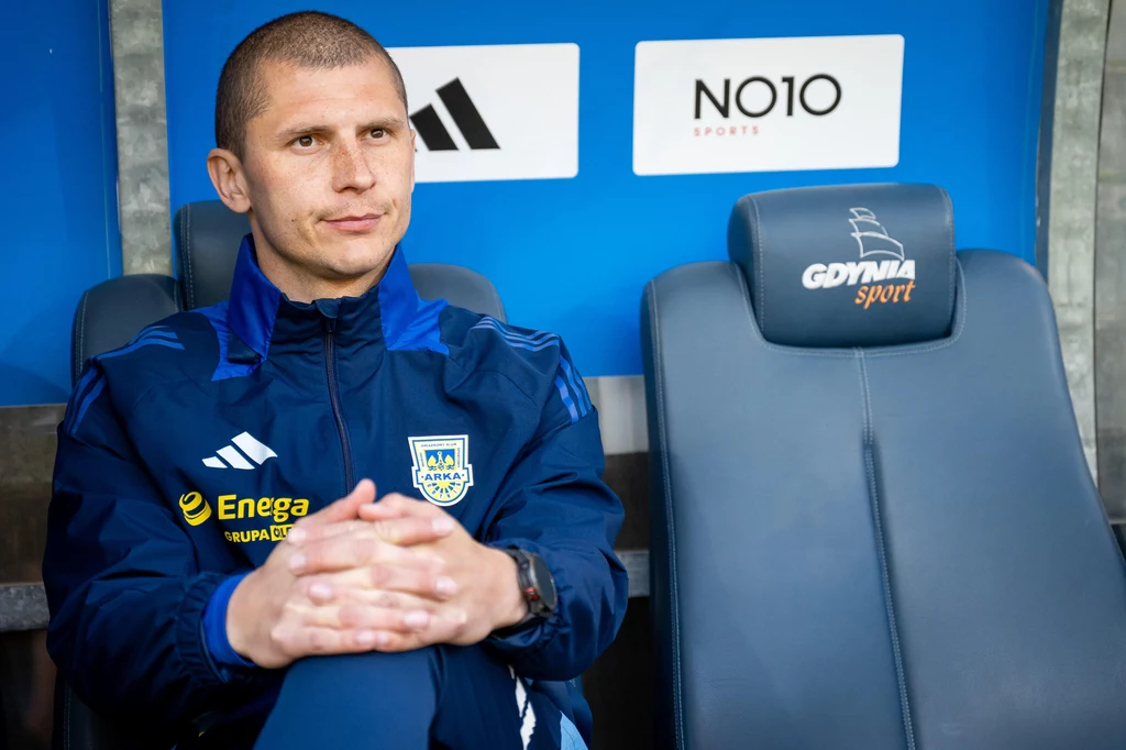 Dawid Szwarga Mężczyzna w sportowym dresie z logotypami sponsorów siedzi na stadionowej ławce, w tle widoczne niebieskie elementy oraz napisy związane z klubem i sponsorem.