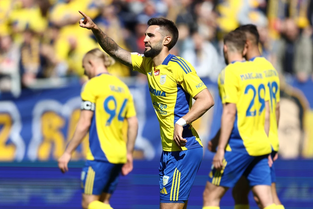 Arka Gdynia Piłkarz w żółto-niebieskim stroju wskazuje ręką kierunek podczas meczu, wokół niego kilku innych zawodników tej samej drużyny, w tle rozmazani kibice na trybunach.