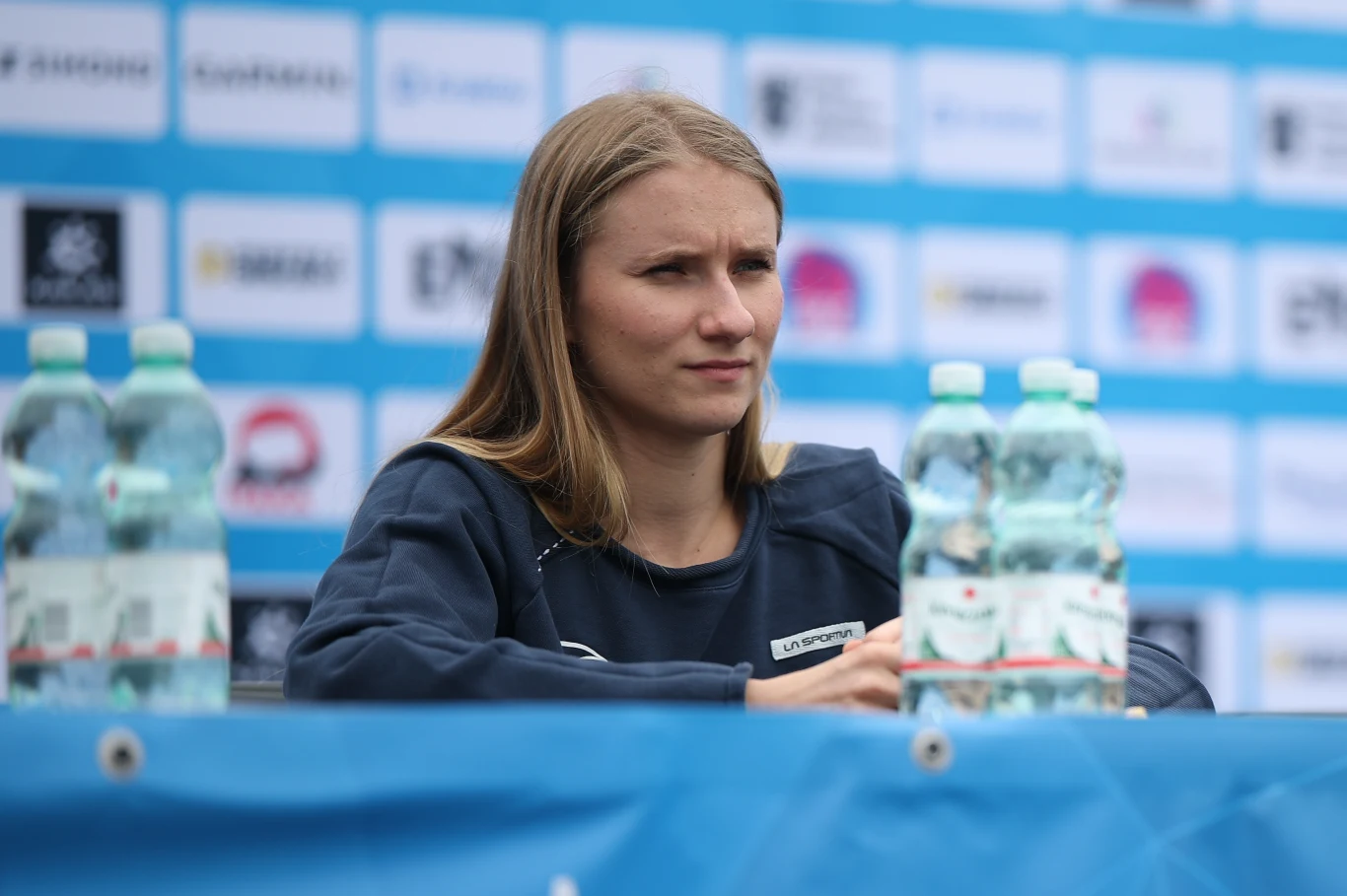 Aleksandra Kałucka Młoda kobieta w sportowym stroju siedzi przy stole podczas konferencji prasowej lub spotkania, na stole znajdują się butelki z wodą mineralną, w tle widoczne są plansze z logotypami sponsorów.