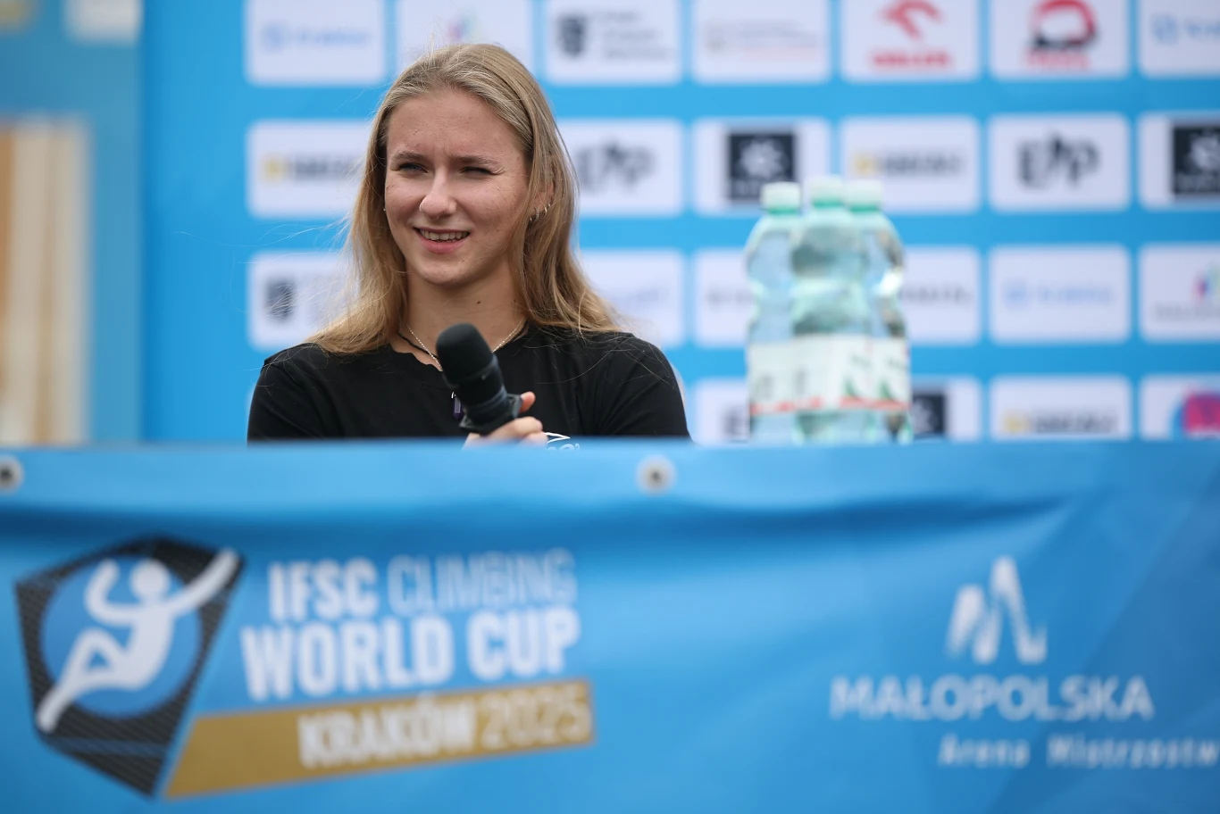 Natalia Kałucka Młoda kobieta z mikrofonem siedzi przy stole podczas konferencji prasowej zawodów wspinaczkowych IFSC Climbing World Cup w Krakowie, w tle widoczne logotypy sponsorów oraz butelka wody.