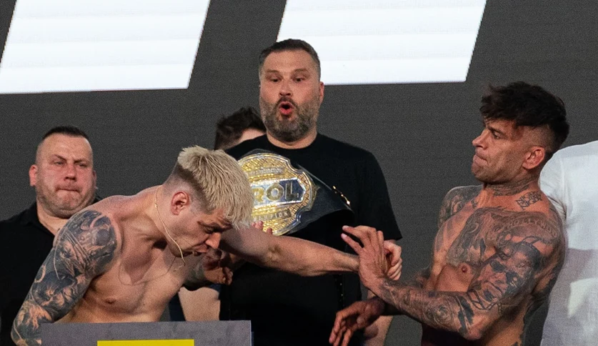 Prime MMA 13: Taazy vs Tańcula Dwóch wytatuowanych mężczyzn podczas konfrontacji twarzą w twarz, jeden z nich uderza drugiego w twarz, w tle osoba z pasem mistrzowskim i dwie inne osoby obserwujące sytuację.