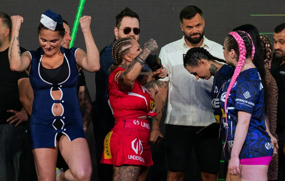 Grupa osób na scenie, w centrum uwagi kobieta w czerwonym stroju sportowym i kobieta w granatowym stroju stylizowanym na kostium, reszta uczestników uśmiechnięta lub skupiona, kolorowe fryzury i tatuaże.