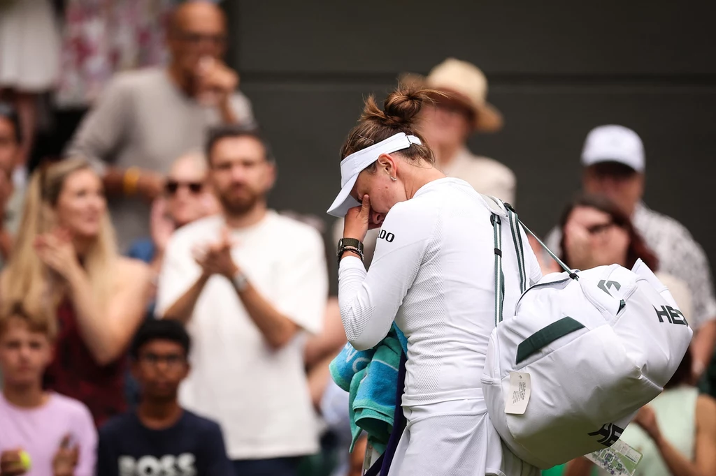 Barbora Krejcikova Zawodniczka tenisa ubrana na biało opuszcza kort z rakietą i torbą sportową, ocierając łzę z twarzy, w tle widać kibiców obserwujących i bijących brawo.