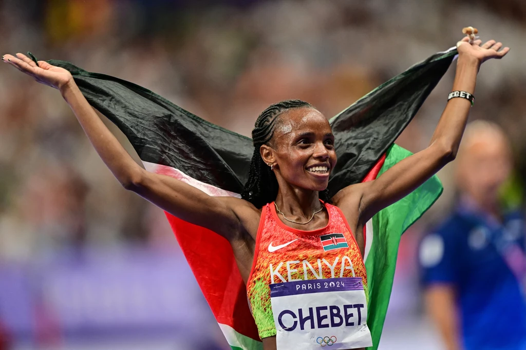 Beatrice Chebet Kenijska lekkoatletka cieszy się ze zwycięstwa, trzymając flagę swojego kraju szeroko rozwiniętą nad głową, uśmiechnięta, w stroju sportowym z nazwiskiem i nazwą kraju na koszulce, na tle rozmytej publiczności.
