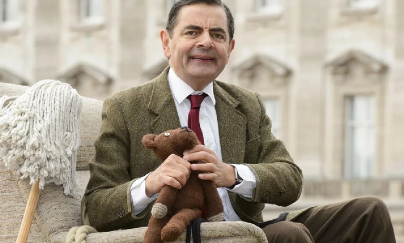 Rowan Atkinson Mężczyzna w marynarce i czerwonym krawacie siedzi na fotelu, trzymając pluszowego misia w dłoniach, obok widoczny mop, w tle rozmyty budynek o klasycznej architekturze.