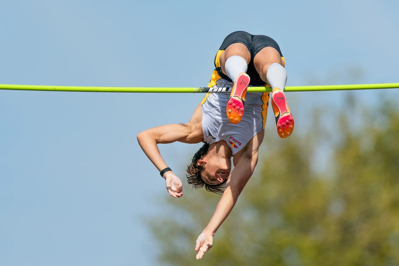 Sportowiec podczas skoku o tyczce w powietrzu, jego ciało przechodzi nad poprzeczką, wyraźnie widoczne dynamiczne napięcie mięśni oraz kolorowe obuwie z kolcami, tło rozmyte, niebo i drzewa.