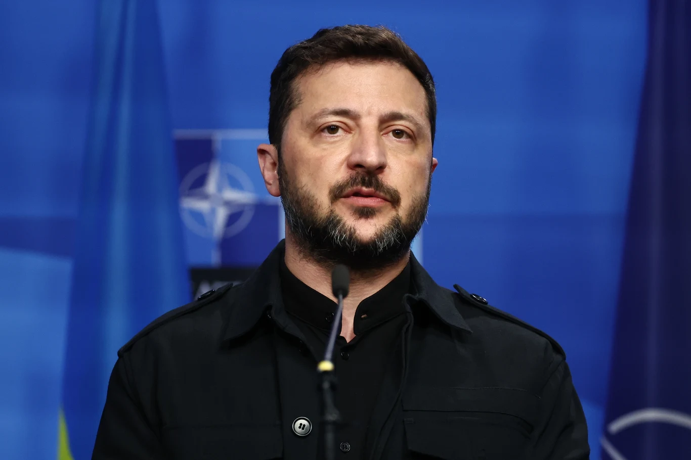 Wołodymyr Zełenski odbył rozmowę z Donaldem Trumpem Mężczyzna z brodą i ciemnymi włosami przemawia przy mikrofonie na tle flag Ukrainy i NATO.