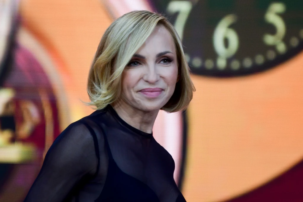 Anna Wyszkoni Kobieta o blond włosach w półformalnym, ciemnym stroju na tle kolorowego zegara.