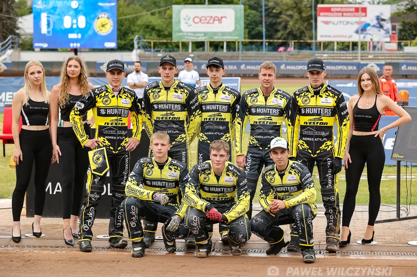 Grupa zawodników żużlowych w żółto-czarnych kombinezonach ustawionych do wspólnej fotografii na stadionie, w towarzystwie czterech kobiet ubranych w czarne stroje sportowe, w tle widoczne trybuny oraz reklamy.