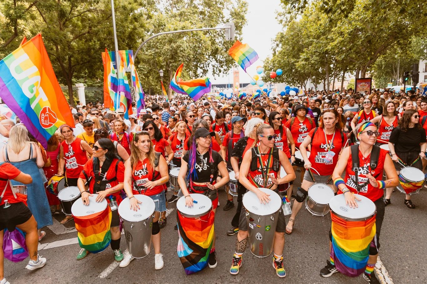 Ulicami Madrytu przeszła największa w Europie parada równości Grupa ludzi maszeruje ulicą podczas parady równości, uczestnicy ubrani w kolorowe stroje i opaski, niektórzy grają na bębnach, widoczne liczne flagi w barwach tęczowych, dookoła tłum i dużo zieleni.