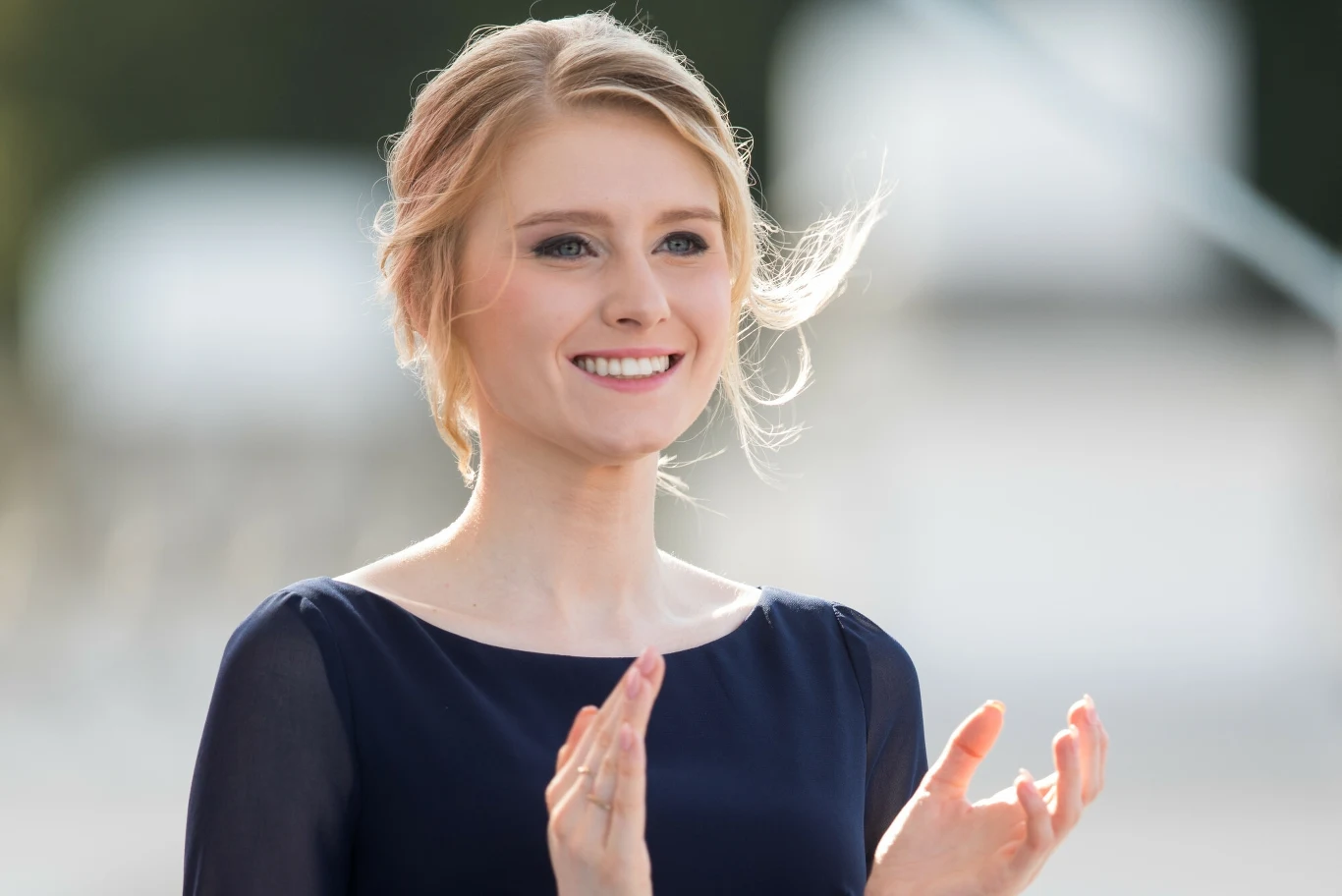 Młoda kobieta z jasnymi włosami upiętymi w kok, uśmiechnięta, ubrana w elegancką, granatową sukienkę, klaszcze w dłonie. W tle rozmyte jasne elementy, naturalne światło.
