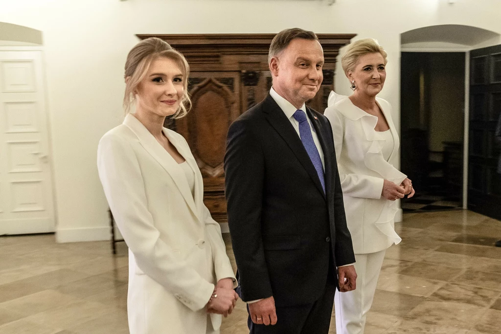 Kinga Duda z rodzicami Trzy elegancko ubrane osoby w jasnych garniturach stoją razem w przestronnym, jasnym wnętrzu z drewnianą szafą w tle, wszystkie spoglądają w stronę aparatu i subtelnie się uśmiechają.