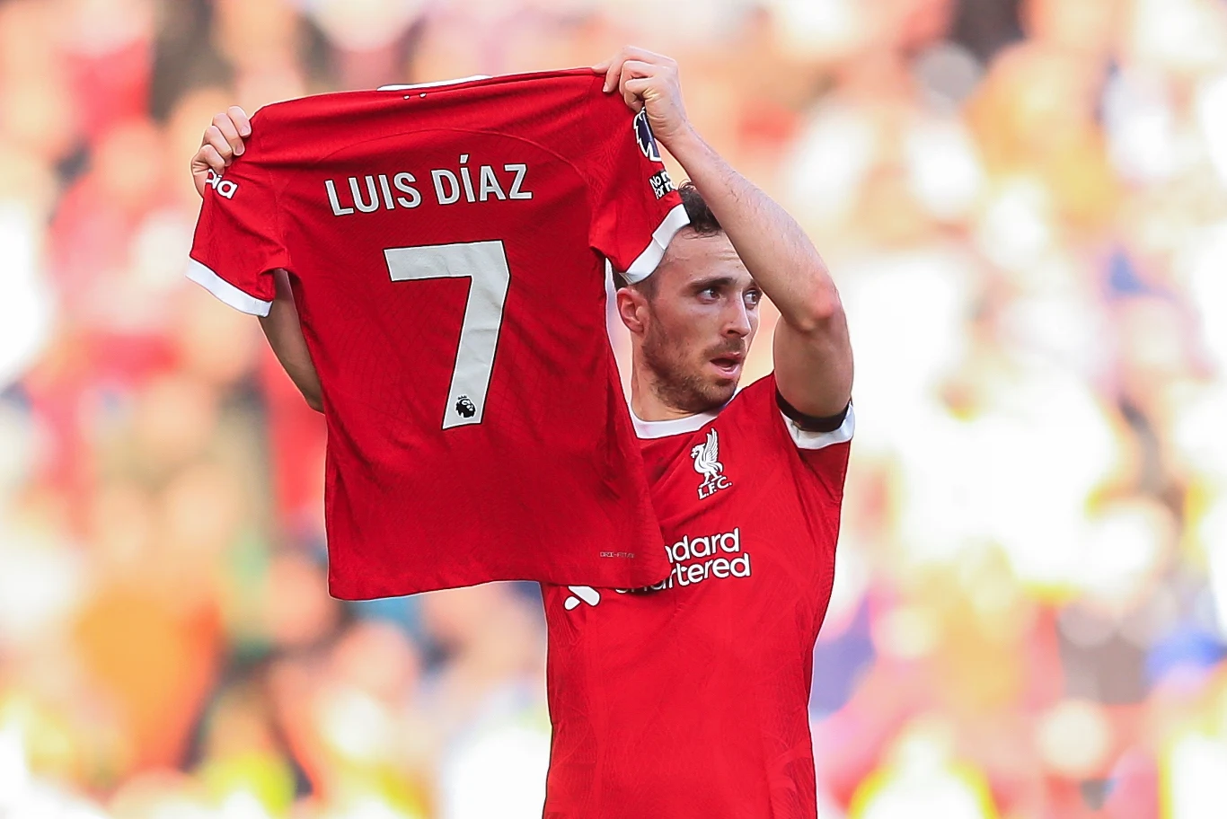 Piłkarz ubrany w czerwony strój Liverpoolu unosi koszulkę z napisem LUIS DÍAZ i numerem 7 na tle rozmytego stadionu pełnego kibiców.