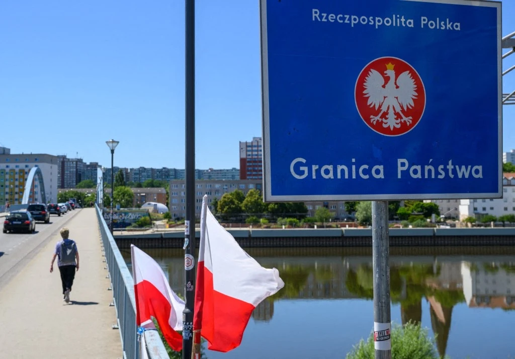 Polska wprowadza kontrole na granicy z Niemcami i Litwą Tablica z godłem Polski i napisem Granica Państwa umieszczona przy rzece, obok niej dwie biało-czerwone flagi. W tle most, przechodzień i samochody, a dalej zabudowa miejska.