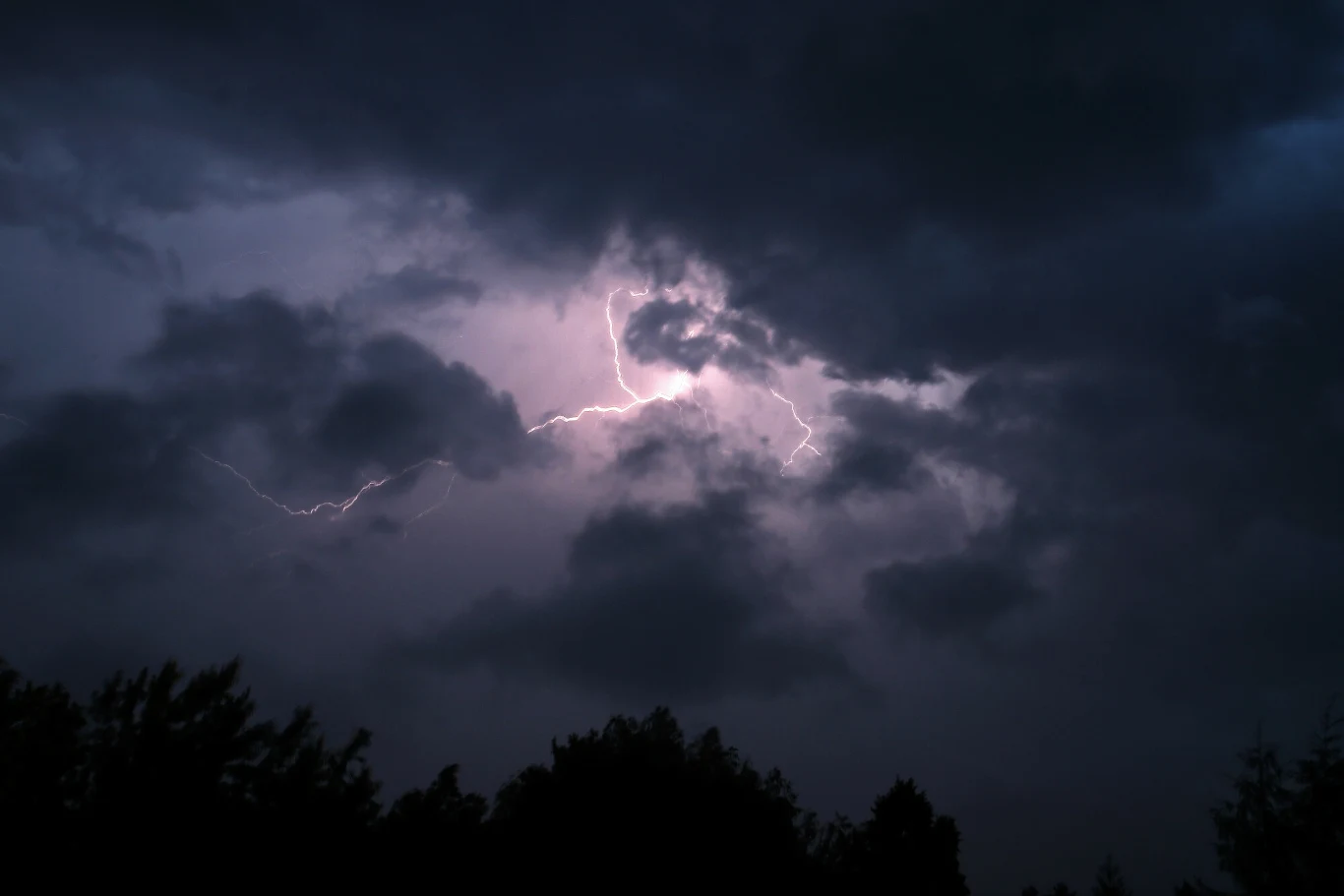 W nocy z niedzieli na poniedziałek na południu Polski mogą wystąpić burze Błyskawica rozświetlająca ciemne, burzowe chmury nad linią drzew.