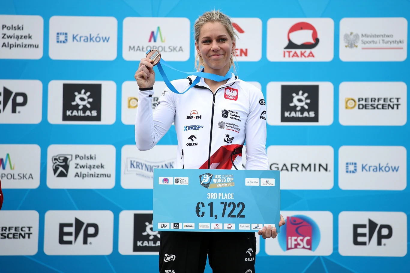 Aleksandra Mirosław sportsmenka w biało-czerwonym stroju z logotypami, trzyma medal oraz tabliczkę z napisem 3rd place i wysokością nagrody pieniężnej, tło stanowią loga sponsorów i organizatorów wydarzenia
