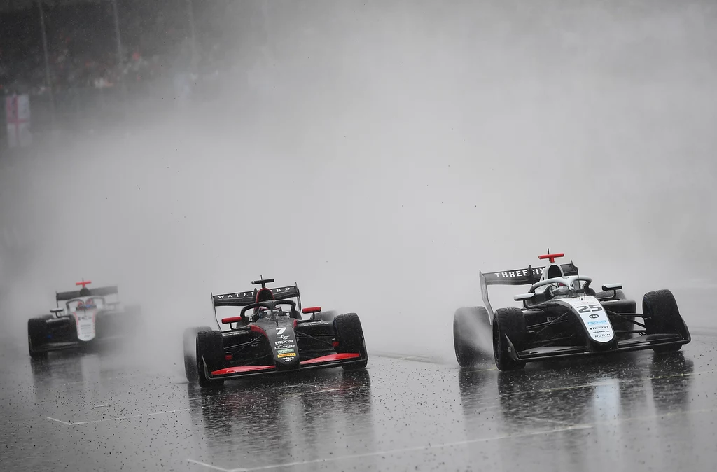 Roman Biliński (#25) 4. podczas deszczowej rywalizacji na torze Silverstone Trzy bolidy wyścigowe jadą po mokrym torze wyścigowym podczas intensywnego deszczu, podnosząc za sobą gęstą chmurę wody ograniczającą widoczność.