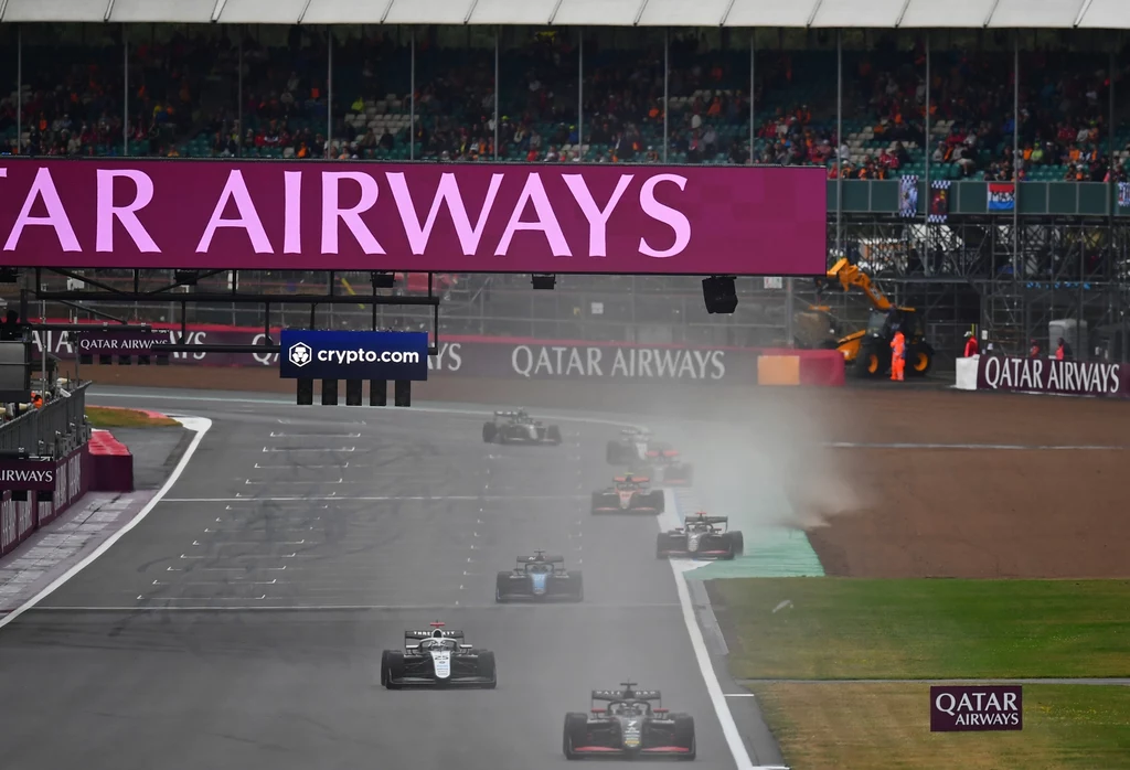 Wyścig Formuły 3 na torze Silverstone Wyścigowe bolidy Formuły 1 poruszające się po torze w warunkach mokrej nawierzchni, unosząc za sobą chmurę wody, nad torem widoczny baner reklamowy Qatar Airways, a w tle trybuny z publicznością.