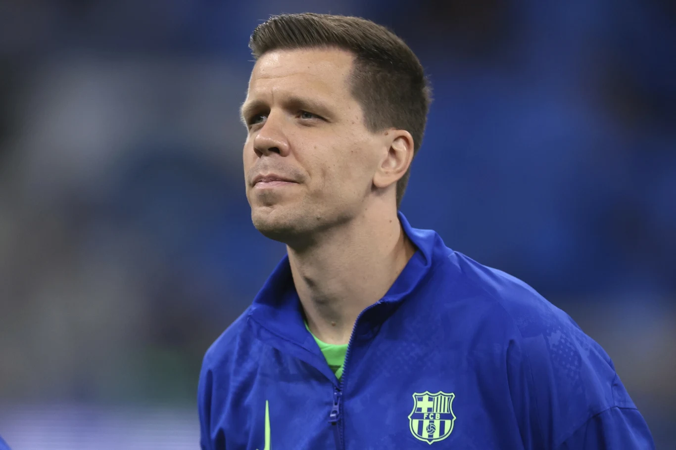 Wojciech Szczęsny Mężczyzna w niebieskiej bluzie sportowej z herbem klubu piłkarskiego na piersi, patrzący w dal, w tle rozmazane niebieskie światło stadionu.