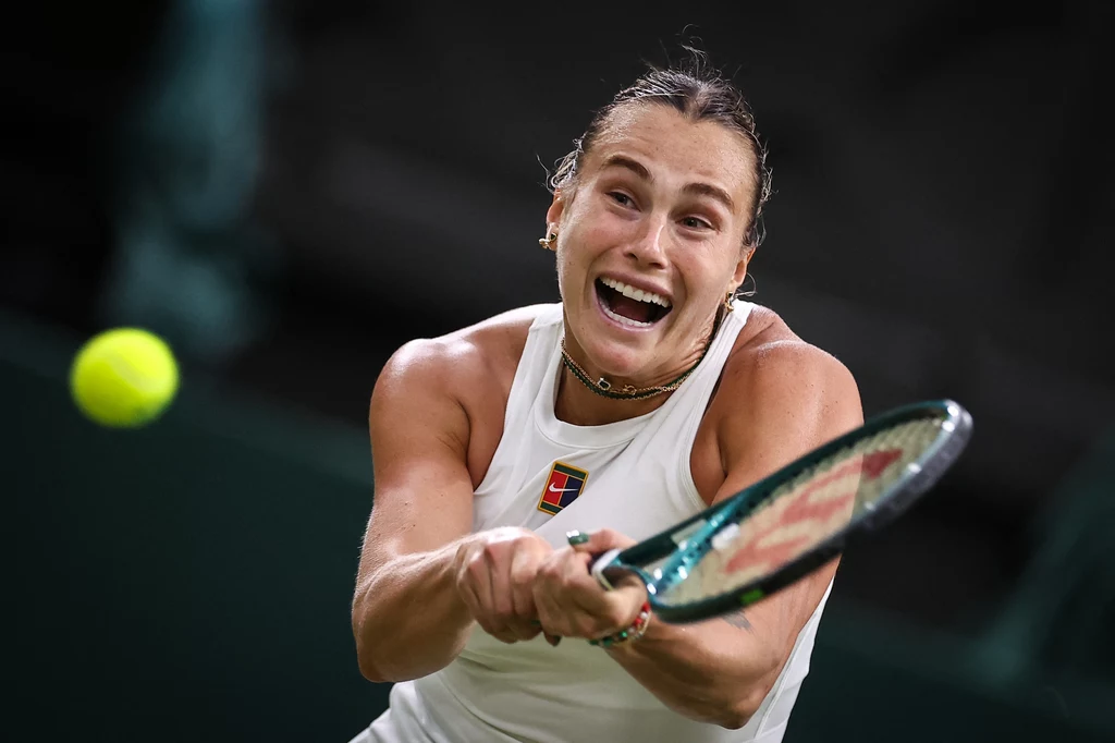 Zawodniczka w białym stroju sportowym uderza piłkę tenisową rakietą, dynamiczna scena rozgrywa się na tle nieostrego boiska, twarz zawodniczki wyraża skupienie i determinację.