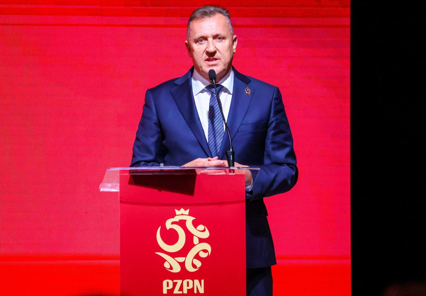 Cezary Kuelsza Mężczyzna w granatowym garniturze wygłasza przemówienie przy mównicy z logo Polskiego Związku Piłki Nożnej na czerwonym tle.