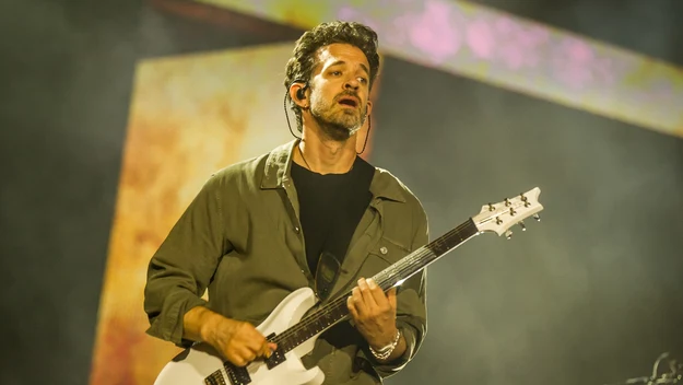 Mężczyzna w zielonej koszuli grający na białej gitarze elektrycznej podczas występu na scenie, w tle oświetlenie sceniczne i mgła.