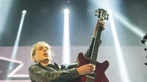 Muzyk grający na gitarze basowej podczas koncertu, dynamiczna poza podkreślająca energię występu, artysta ubrany w skórzaną kurtkę i jeansy, kolorowe światła sceniczne oraz widoczny sprzęt muzyczny dopełniają atmosfery koncertowej.