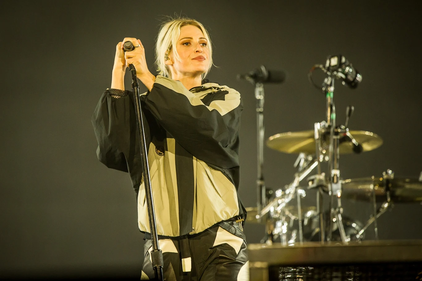 Open'er Festival 2025: Linkin Park Kobieta na scenie trzyma mikrofon, ubrana w luźną, kontrastującą kurtkę, w tle perkusja i mikrofony sugerujące występ muzyczny.