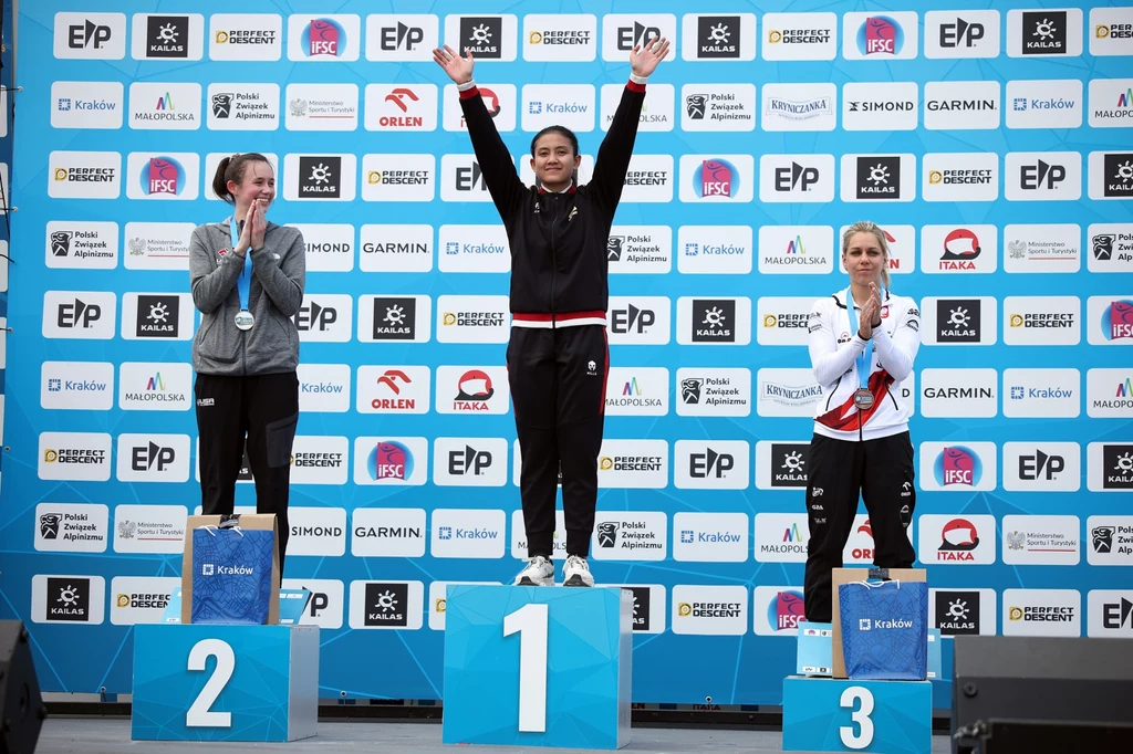 Podium zawodów PŚ kobiet we wspinaczce w Krakowie. Od lewej: Emma Hunt, Desak Made Rita Kusuma Dewi i Aleksandra Mirosław Trzy kobiety stoją na podium podczas dekoracji zwycięzców zawodów sportowych, środkowa zawodniczka podnosi ręce w geście triumfu, pozostałe dwie klaskają.
