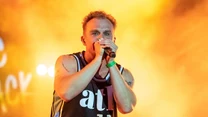 Mężczyzna podczas występu muzycznego na scenie, ubrany w sportową koszulkę z numerem 11, trzyma mikrofon i śpiewa, w tle intensywne, żółto-pomarańczowe światło sceniczne.