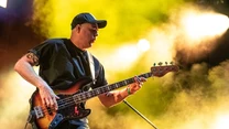 Muzyk grający na gitarze basowej na scenie, dynamiczne oświetlenie w tle tworzy energiczną atmosferę koncertu, artysta ubrany w czarną koszulkę i czapkę z daszkiem.