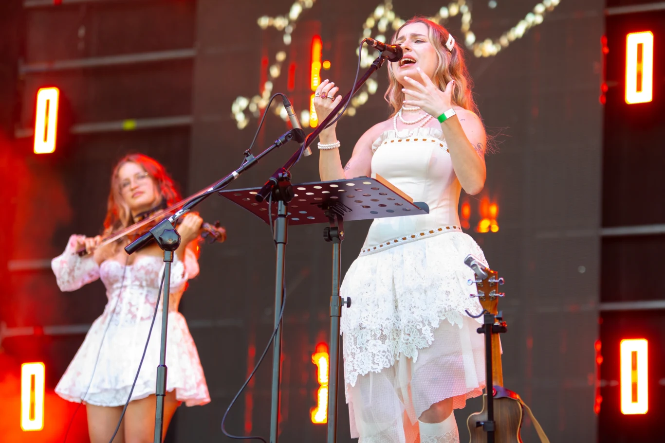 Lor Dwie kobiety ubrane w białe kostiumy sceniczne występują na scenie, jedna śpiewa do mikrofonu, druga gra na skrzypcach, w tle widoczne kolorowe światła sceniczne i dekoracje.