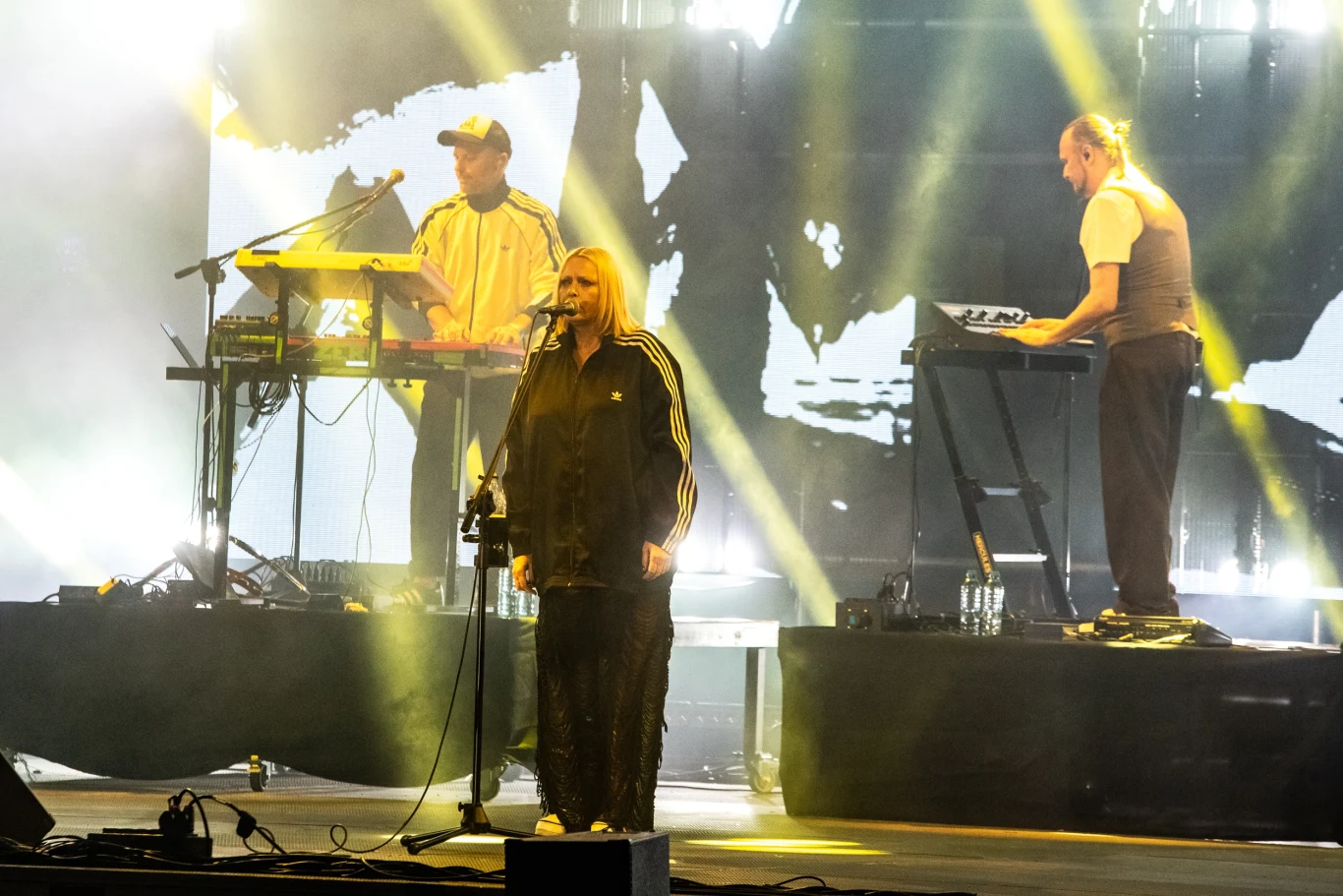 Trzech muzyków na scenie podczas koncertu, w centrum kobieta śpiewająca do mikrofonu, po jej bokach dwaj mężczyźni obsługujący instrumenty klawiszowe, w tle intensywne światła sceniczne i wizualizacje