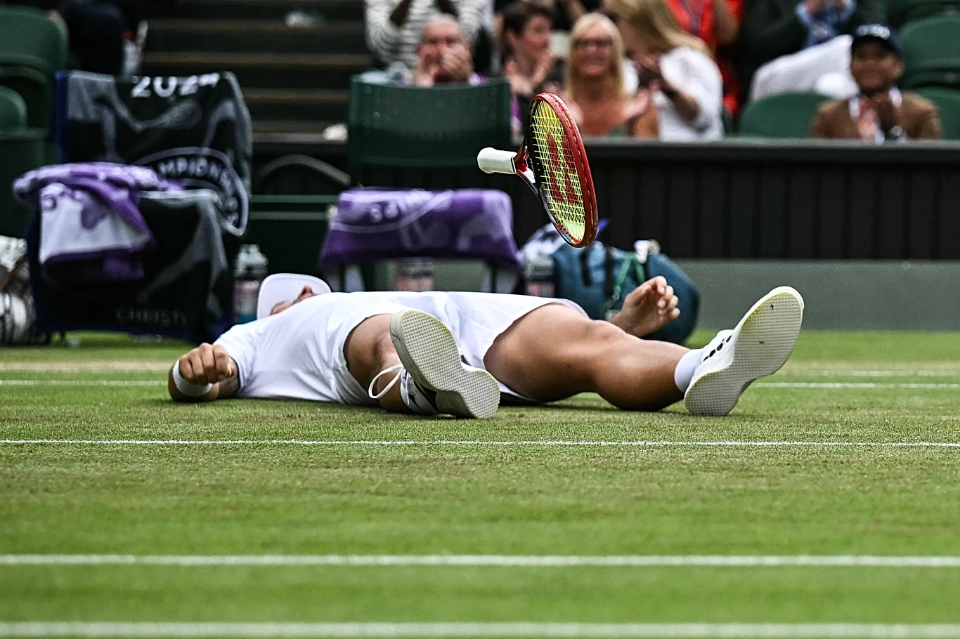 Radość Jana Zielińskiego na kortach Wimbledonu Tenisista leżący na murawie kortu, prawdopodobnie celebruje wygraną lub moment zakończenia meczu, nad nim unosi się jeszcze rakieta wypuszczona z ręki, w tle publiczność oraz trybuny.