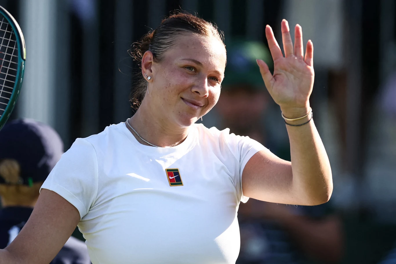 Amanda Anisimova Tenisistka ubrana w białą koszulkę unosi rękę, machając do widowni, z rakietą tenisową trzymaną w tle. Twarz kobiety jest uśmiechnięta, a na szyi ma sportowy wisiorek.