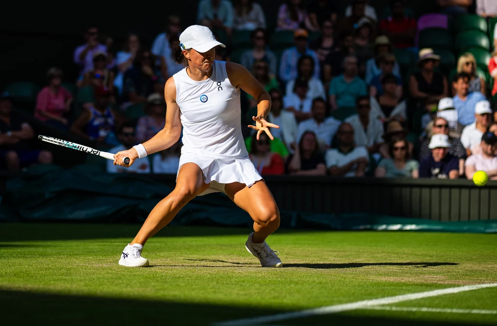 Iga Świątek, Wimbledon 2023. Mecz z Belindą Bencic Tenisistka w białym stroju sportowym wykonuje dynamiczny ruch podczas meczu na trawiastym korcie, w tle widoczna publiczność.