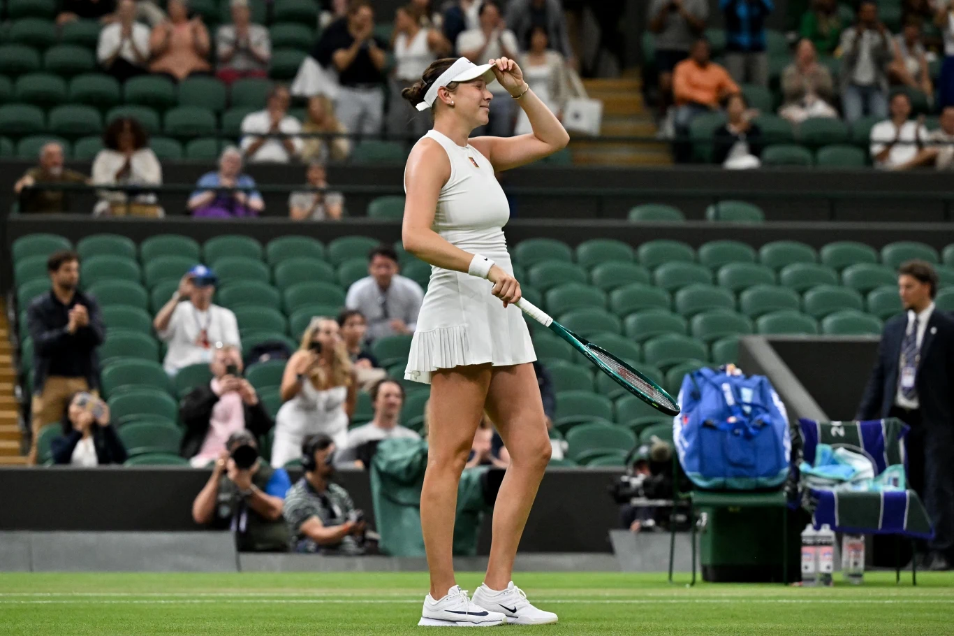 Tenisistka w białym stroju sportowym na korcie, trzymająca rakietę i poprawiająca czapkę, w tle trybuny z widownią oraz sprzęt sportowy.
