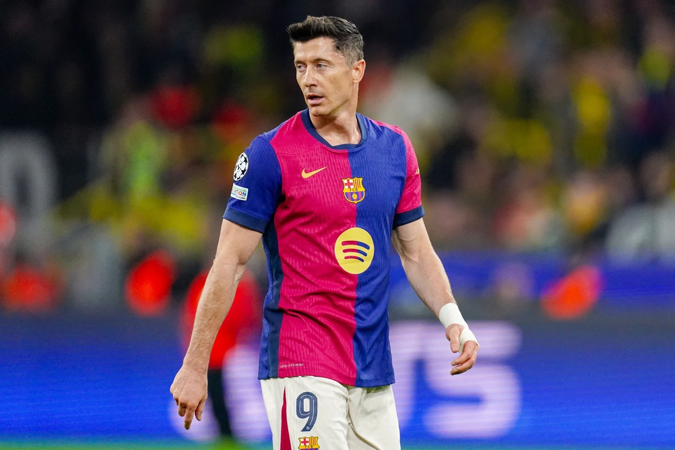 Piłkarz w stroju FC Barcelona z numerem 9 na tle rozmytego stadionu podczas meczu piłkarskiego, widoczna opaska na nadgarstku oraz sponsor na koszulce.