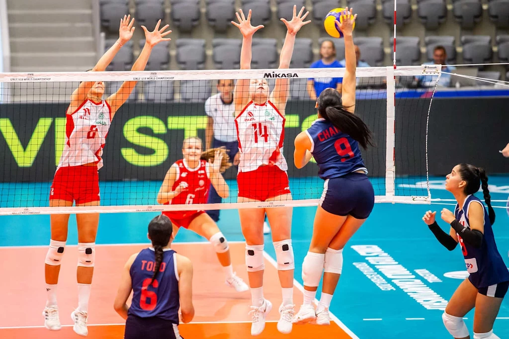 Mecz Polska - Peru na mistrzostwach świata do lat 19 Zawodniczki dwóch drużyn siatkarskich w trakcie dynamicznej akcji na boisku, jedna z zawodniczek z drużyny w granatowych strojach wykonuje atak na siatce, podczas gdy trzy zawodniczki w biało-czerwonych strojach próbują zablokować piłkę