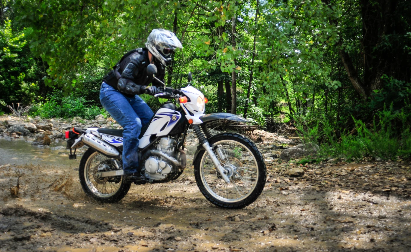 Motocykl Yamaha XT250 rocznik 2008. Wersja wojskowa dostanie m.in. specjalne malowanie i uchwyt na narty (zdj. poglądowe) Motocyklista w pełnym stroju ochronnym, w tym kasku i kurtce, prowadzi biały motocykl terenowy na błotnistej i wilgotnej ścieżce w lesie, otoczony intensywną zielenią drzew.