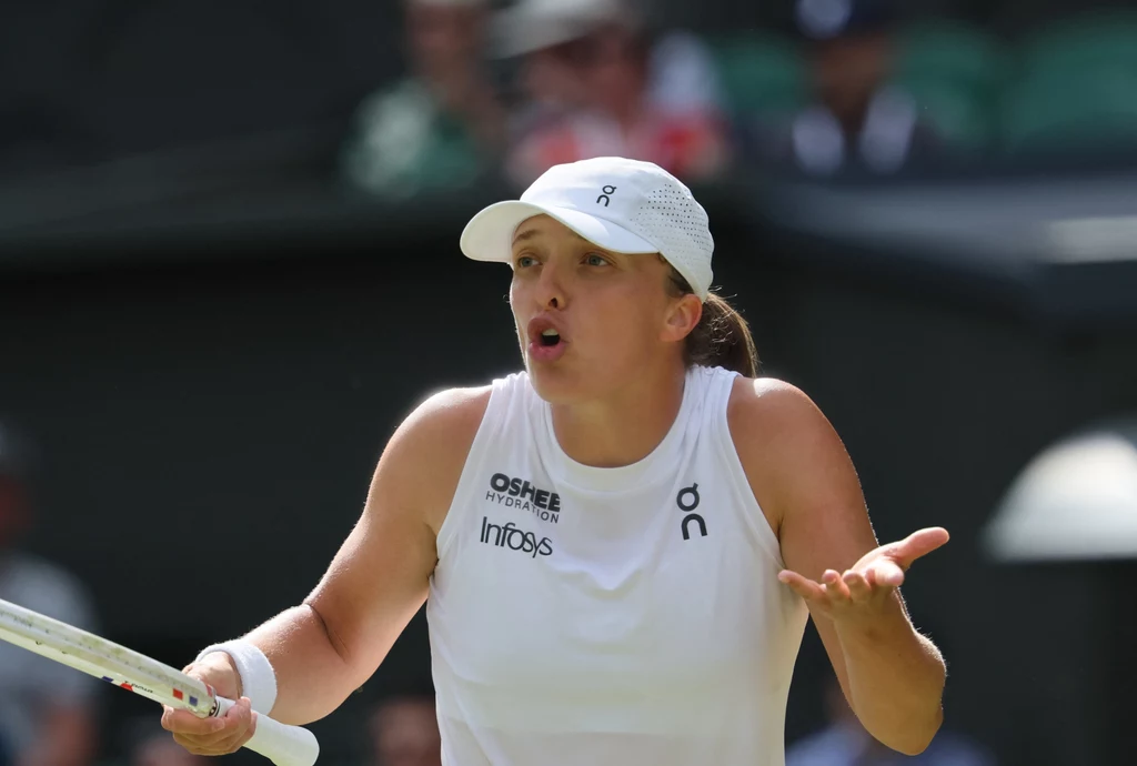 Iga Świątek Zawodniczka tenisowa w białym stroju sportowym z rakietą w ręku, w widocznym geście emocji lub frustracji podczas meczu, z zaciśniętymi ustami i rozłożonymi rękami, na tle rozmytej widowni.