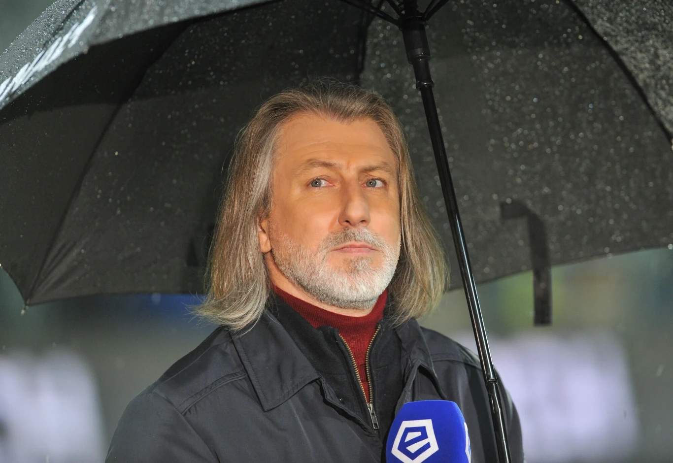 Mężczyzna o siwych włosach stojący pod czarnym parasolem w trakcie deszczu, trzymający mikrofon z logo stacji sportowej, ubrany w ciemną kurtkę i bordowy golf.
