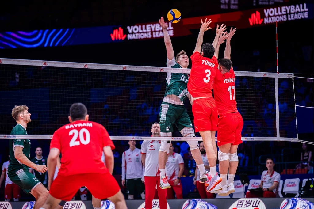 Liga Narodów siatkarzy. Turcja w tym sezonie przegrała m.in. z Polską Siatkarz w zielonym stroju wykonuje atak na piłce nad siatką, dwóch zawodników w czerwonych strojach próbuje go zablokować, w tle widać innych graczy oraz sędziów, na tablicach widoczne logo Volleyball Nations League.