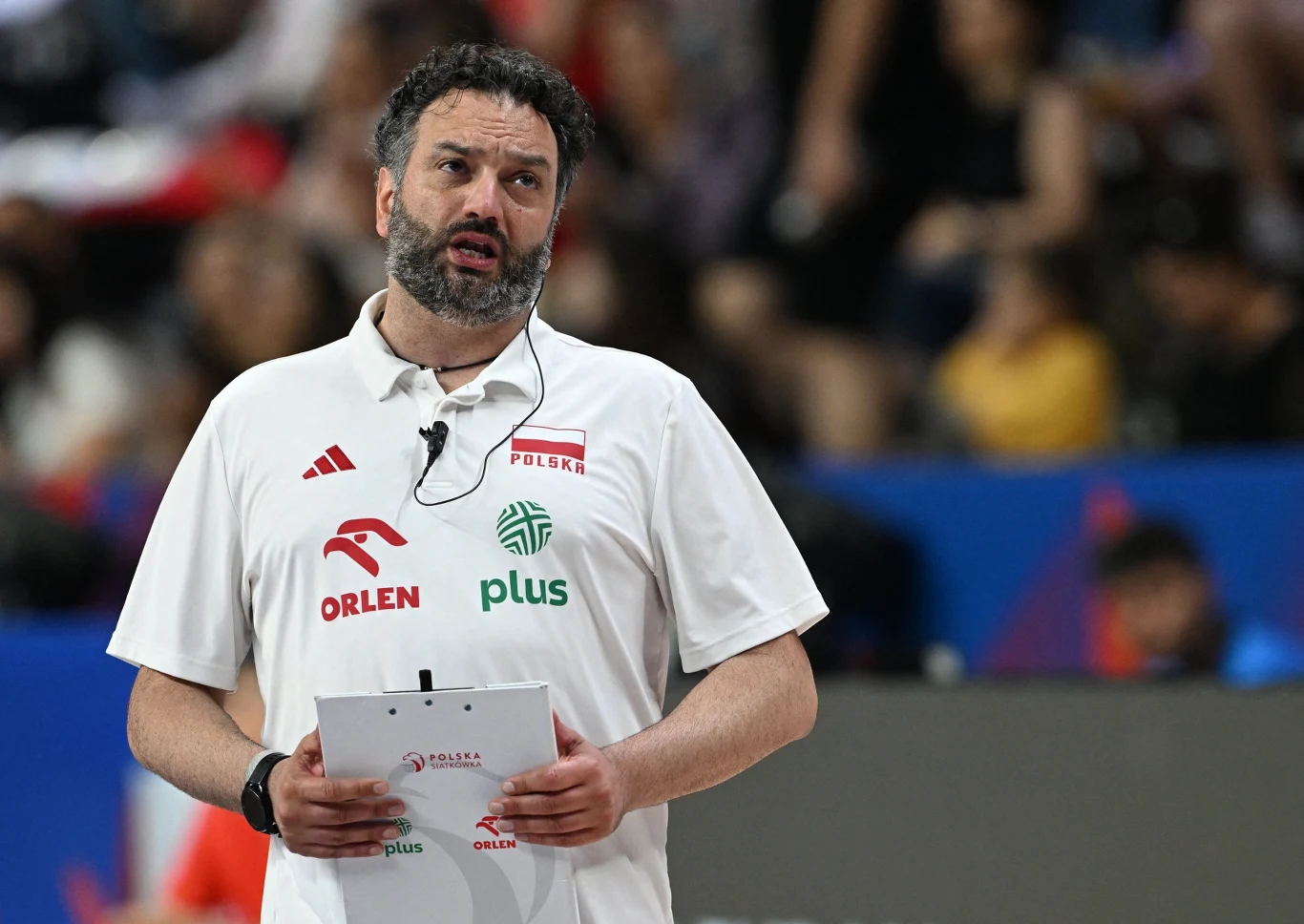 Stefano Lavarini Trener w białej koszulce z emblematami sponsorów oraz napisem Polska, trzymający notes i zestaw słuchawkowy z mikrofonem, na tle rozmazanej publiczności sportowej.
