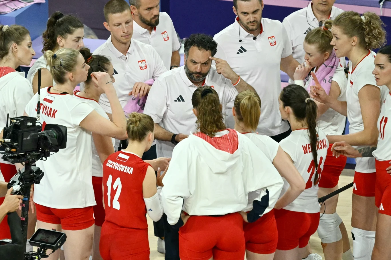 Zespół siatkarek w biało-czerwonych strojach wraz z trenerem omawia strategię podczas przerwy w meczu, wokół stoją członkowie sztabu i ekipa techniczna.