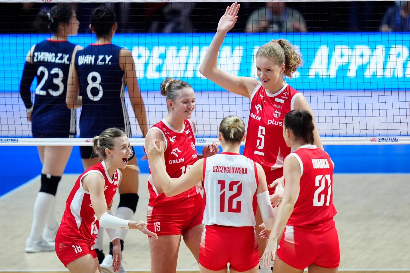 Grupa polskich siatkarek ubranych w czerwone stroje cieszy się po zdobyciu punktu na boisku, za nimi widoczne dwie zawodniczki drużyny przeciwnej w granatowych koszulkach