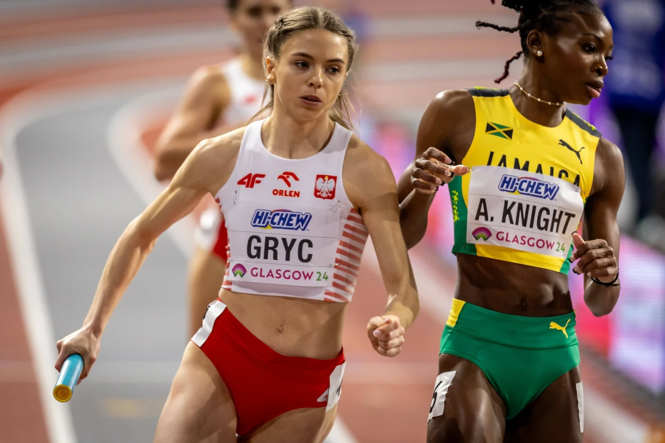 Zawodniczka polskiej sztafety biegnie z pałeczką podczas zawodów lekkoatletycznych, obok rywalka z Jamajki, obie ubrane w stroje sportowe w barwach narodowych, na tle bieżni stadionowej.