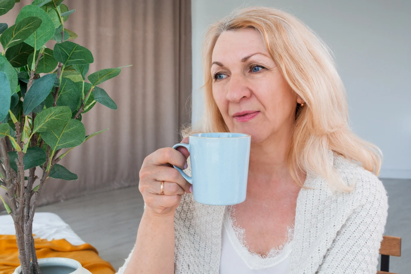 Na co pomaga herbata gryczana? Starsza kobieta o blond włosach trzyma w dłoni niebieski kubek i siedzi przy stole, obok rośliny w doniczce, w jasnym, spokojnym wnętrzu.