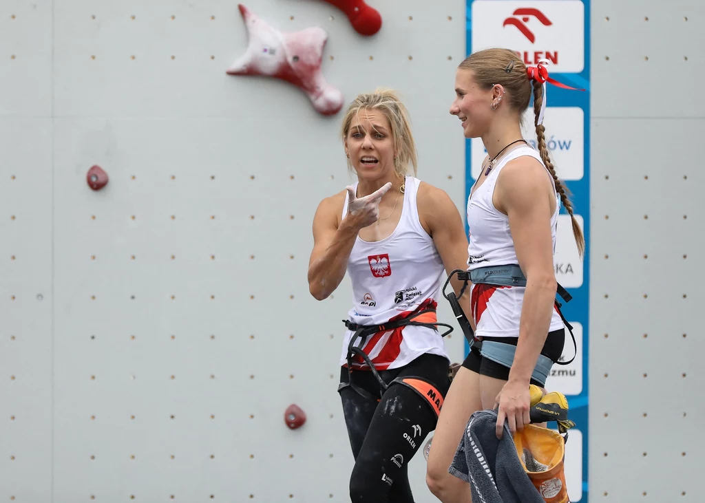 Aleksandra Mirosław (po lewej) i Natalia Kałucka Dwie kobiety w sportowych uprzężach i koszulkach z polskim godłem stoją przy ściance wspinaczkowej, jedna z nich wyraża silne emocje, druga trzyma woreczek z magnezją i uśmiecha się.