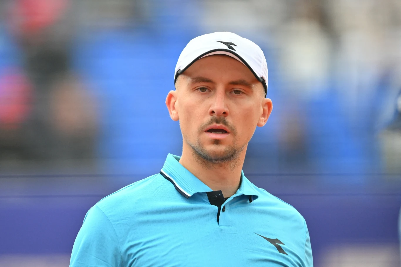 Jan Zieliński Mężczyzna w sportowej czapce i błękitnej koszulce polo stoi na tle rozmytych trybun stadionu, patrząc poważnie przed siebie.